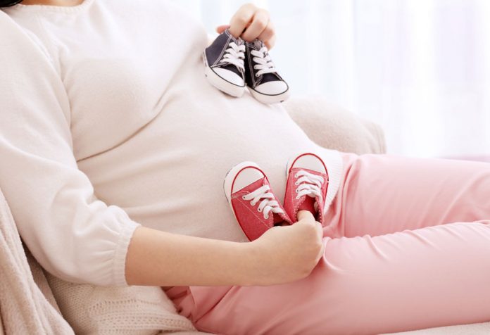 Pregnant woman holding two pairs of shoes (red and blue) close to her belly